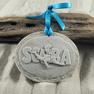 Scuba Diver Ornament made with real sand depicting raised large lettering that say SCUBA and overlayed is a diver with bubbles coming from his tank.  The ornament is oval with a edged board. Lightly painted to bring out details, glittered and tied with a satin turquoise ribbon and bow. Attached is a tag indicating that it is handcrafted with sand and made in the USA.