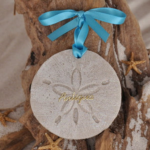 Antiqua sand dollar ornament.  Sand dollar shape, made with sand, that has been whitewashed and glittered.  In the center in gold scripted letters is Antiqua. Tied with a turquoise ribbon and tag that says it is Handcrafted with Sand.
